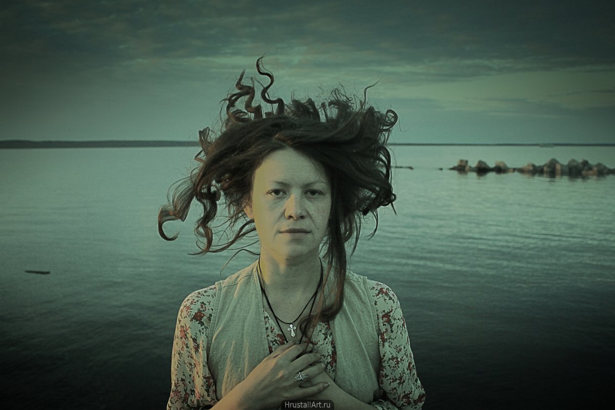 Фотография, портрет молодой женщины с парящими в хаосе волосами, она пристально смотрит на зрителя тяжёлым взглядом, руки прижаты к груди, на шее маленький крестик.