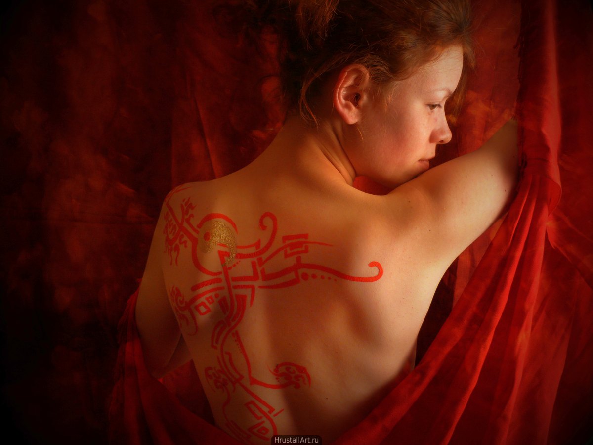 Фотография, девушка с красной росписью на спине полуобернулась к зрителю. Красная ткань и языки пламени.