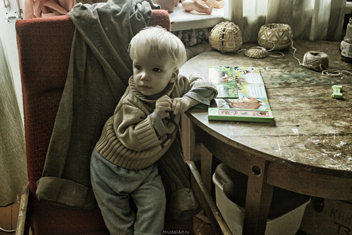 Маленький но очень серъёзный ребёнок в тёплом свитере опирается на винтажный круглый стол. Гранж.